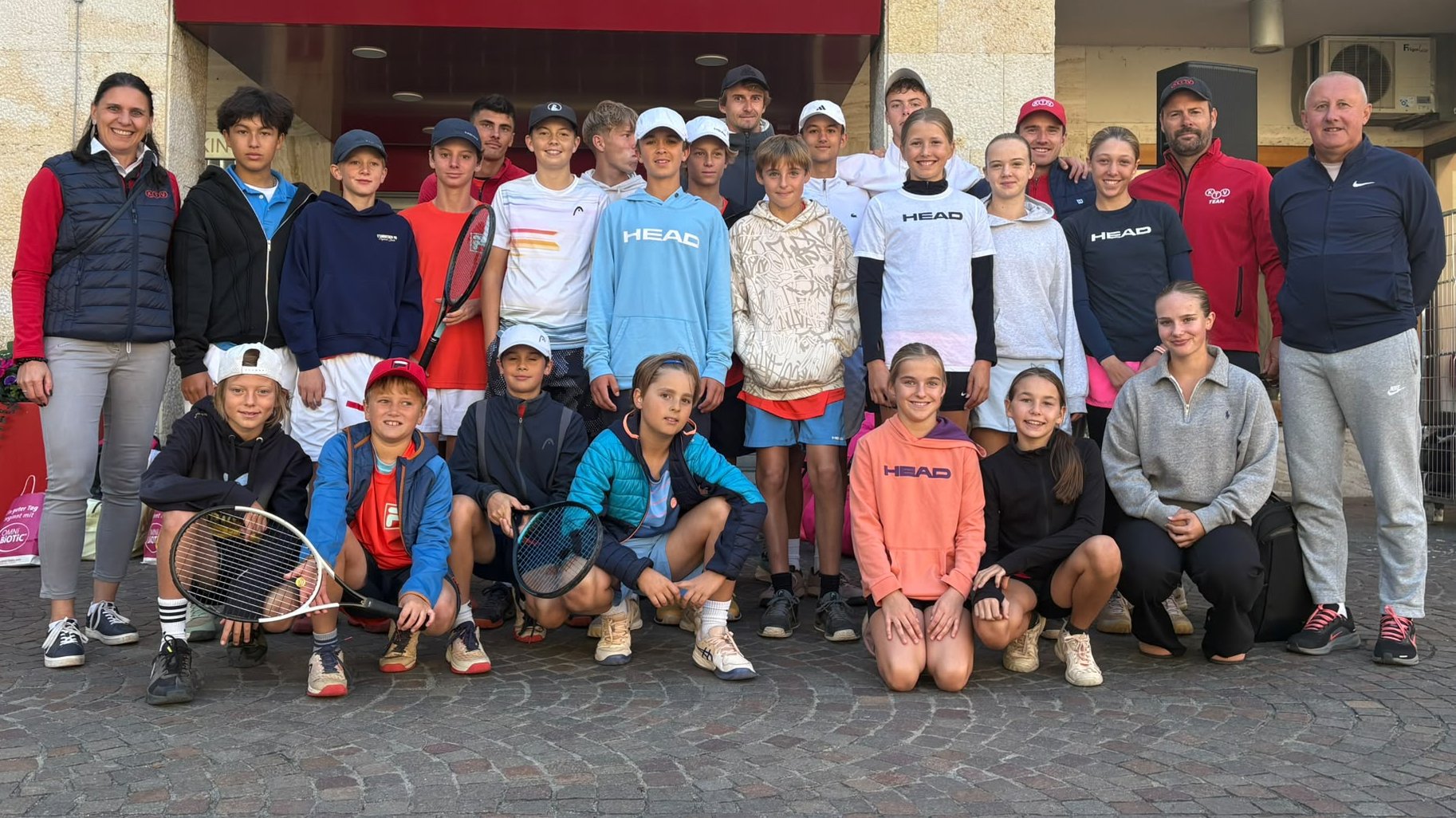 Tennis-Jugendteam mit Betreuern posiert für ein Gruppenfoto vor einem Gebäude.